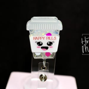 May include: A white and clear badge reel featuring a cartoon coffee cup design. The cup reads "HAPPY PILLS" in pink text and has a smiling face. The reel is filled with glitter and pink accents.