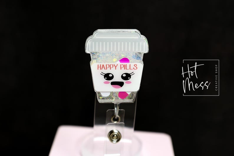 May include: A white and clear badge reel featuring a cartoon coffee cup design. The cup reads "HAPPY PILLS" in pink text and has a smiling face. The reel is filled with glitter and pink accents.