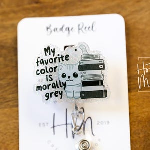 May include: A badge reel with a clear plastic charm featuring a cartoon cat with a stack of books. The charm has the text "My favorite color is morally grey".