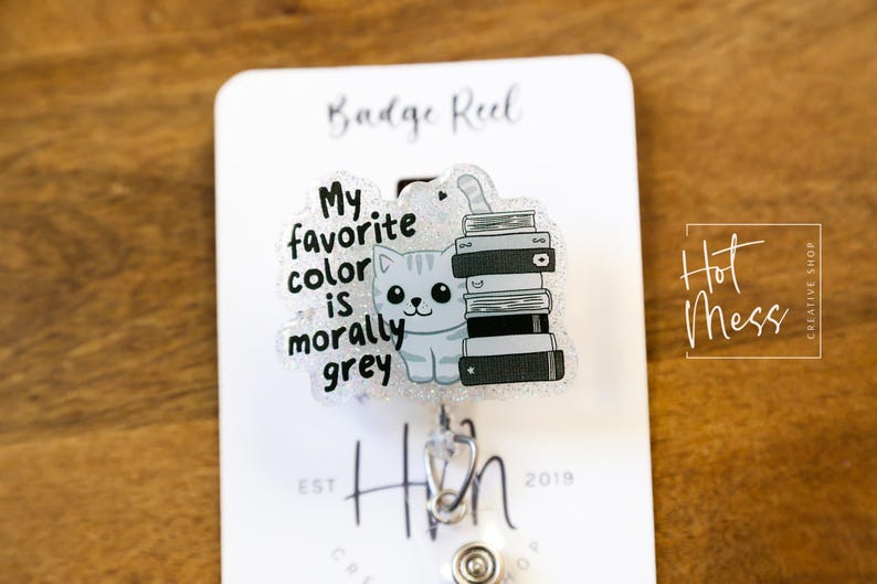 May include: A badge reel with a clear plastic charm featuring a cartoon cat with a stack of books. The charm has the text "My favorite color is morally grey".