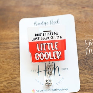 Op de afbeelding: Een witte badge reel met een rood en wit ontwerp met de tekst "DON'T HATE ME JUST BECAUSE I'M A LITTLE COOLER." De reel heeft een metalen clip en een doorzichtige plastic knop. De achtergrond is een houten oppervlak.