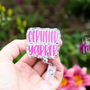 Puede incluir: Un portatarjetas con el texto "CERTIFIED YAPPER" en rosa brillante, contorneado en blanco, sobre un fondo brillante. El portatarjetas se sostiene en una mano, con un fondo floral borroso.