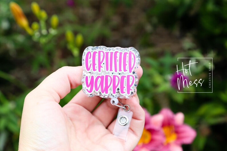 Puede incluir: Un portatarjetas con el texto "CERTIFIED YAPPER" en rosa brillante, contorneado en blanco, sobre un fondo brillante. El portatarjetas se sostiene en una mano, con un fondo floral borroso.