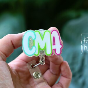 K&ouml;nnte beinhalten: Ein farbenfrohes Ausweis-Jojo mit den Buchstaben "CMA" in Wei&szlig;, umrandet von T&uuml;rkis, Gr&uuml;n und Pink. Das Jojo hat einen transparenten Kunststoffclip und eine einziehbare Kordel. Der Text "Hot Mess Creative Shop" ist im Hintergrund zu sehen.