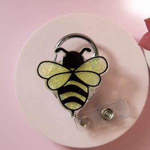 May include: A badge reel with a bee design. The bee has black and yellow stripes with glittery wings. The reel is attached to a silver clip and a clear plastic strap with a snap.