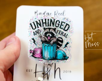 May include: A white glitter badge reel with a black and white raccoon graphic. The raccoon is in a blue trash can and the text "Unhinged and Feral" is above the raccoon. The badge reel is from the shop "Hot Mess Creative Shop."