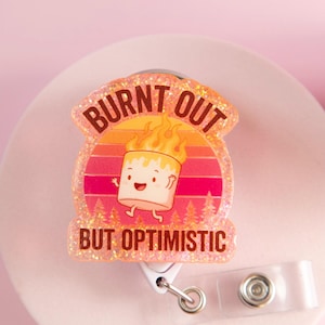 Puede incluir: Un portatarjetas brillante con el texto "BURNT OUT BUT OPTIMISTIC". El diseño presenta una caricatura de malvavisco en llamas sobre un fondo de puesta de sol. El portatarjetas tiene una correa de plástico transparente y un clip de metal.
