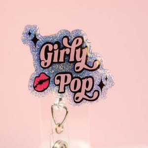 Op de afbeelding: Een glitter badge reel met de woorden "Girly Pop" in roze en zwarte letters, samen met een roze lip-afbeelding. De badge reel is lichtblauw met zilveren glitter en zwarte steraccenten. De achtergrond is effen lichtroze.