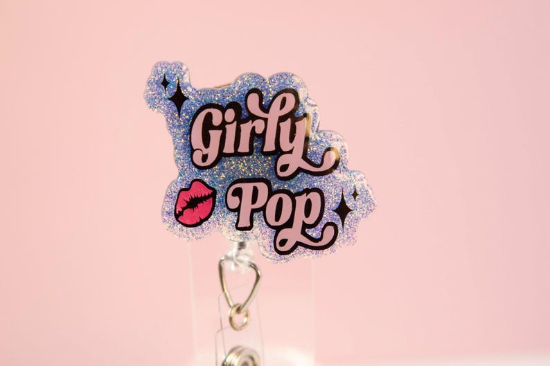 Op de afbeelding: Een glitter badge reel met de woorden "Girly Pop" in roze en zwarte letters, samen met een roze lip-afbeelding. De badge reel is lichtblauw met zilveren glitter en zwarte steraccenten. De achtergrond is effen lichtroze.