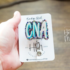 May include: A white card with the text "Badge Reel" and "Hot Mess Creative Shop" printed on it. A blue, white, and purple glitter badge reel with the text "CNA" is attached to the card. The badge reel is attached to a silver retractable badge holder.