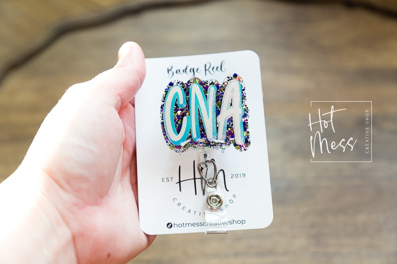 May include: A white card with the text "Badge Reel" and "Hot Mess Creative Shop" printed on it. A blue, white, and purple glitter badge reel with the text "CNA" is attached to the card. The badge reel is attached to a silver retractable badge holder.