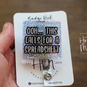 Könnte beinhalten: Ein silberner Glitzer-Badge-Rollenhalter mit dem Text "OOH... THIS CALLS FOR A SPREADSHEET" darauf. Der Badge-Rollenhalter ist an einer weißen Karte mit dem Text "Hot Mess Creative Shop" befestigt.