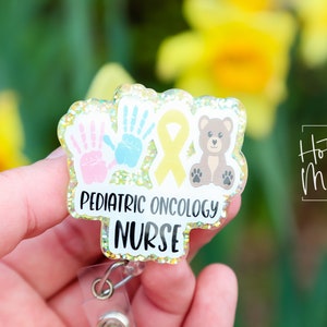 Peut inclure: Un badge jaune paillet&eacute; avec un ruban jaune, deux empreintes de mains et un ours en peluche brun. Le badge indique "Pediatric Oncology Nurse".