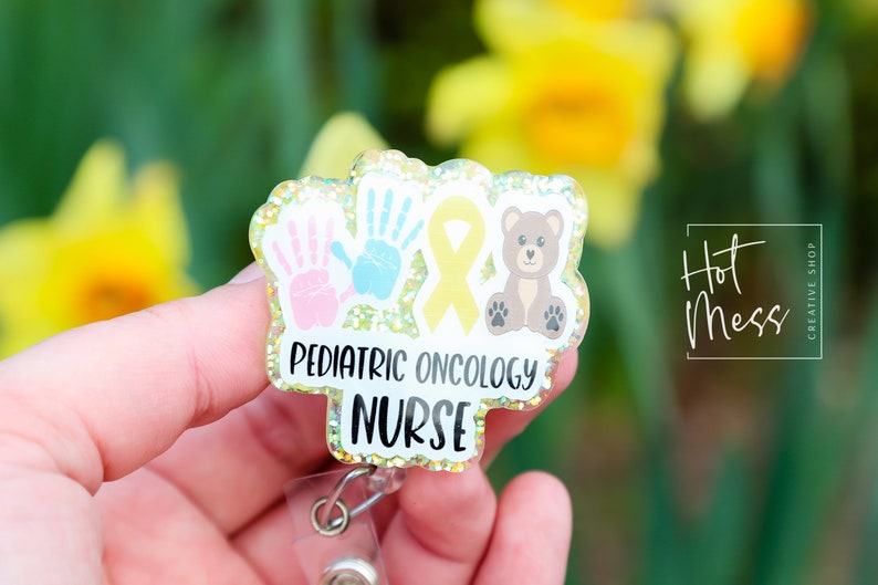 Peut inclure: Un badge jaune paillet&eacute; avec un ruban jaune, deux empreintes de mains et un ours en peluche brun. Le badge indique "Pediatric Oncology Nurse".