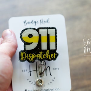 Puede incluir: Un portatarjetas con un dise&ntilde;o de Despachador 911. El dise&ntilde;o presenta el n&uacute;mero 911 en blanco y amarillo, con la palabra "Dispatcher" debajo. El portatarjetas est&aacute; adherido a una tarjeta blanca con las palabras "Badge Reel" en la parte superior.
