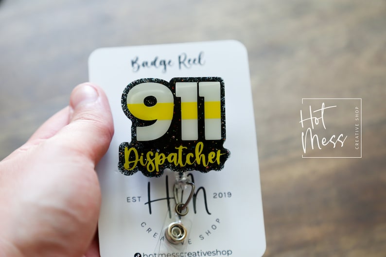 Puede incluir: Un portatarjetas con un dise&ntilde;o de Despachador 911. El dise&ntilde;o presenta el n&uacute;mero 911 en blanco y amarillo, con la palabra "Dispatcher" debajo. El portatarjetas est&aacute; adherido a una tarjeta blanca con las palabras "Badge Reel" en la parte superior.