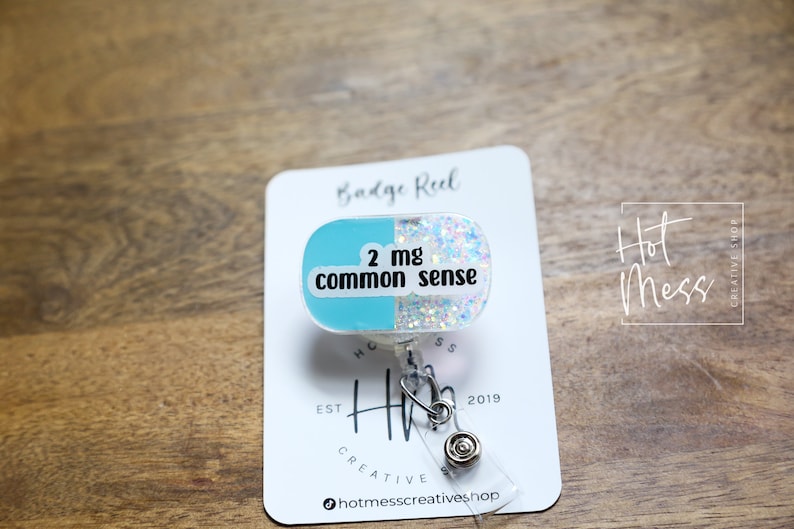 May include: A blue and white glitter badge reel with a pill-shaped charm that says "2 mg common sense".