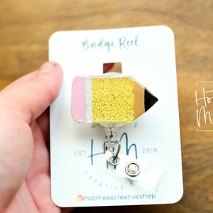 May include: A gold glitter pencil badge reel with a pink eraser and black tip. The reel is attached to a white card with the text "Badge Reel" and "Hot Mess Creative Shop" printed on it.