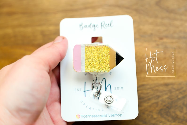 May include: A gold glitter pencil badge reel with a pink eraser and black tip. The reel is attached to a white card with the text "Badge Reel" and "Hot Mess Creative Shop" printed on it.