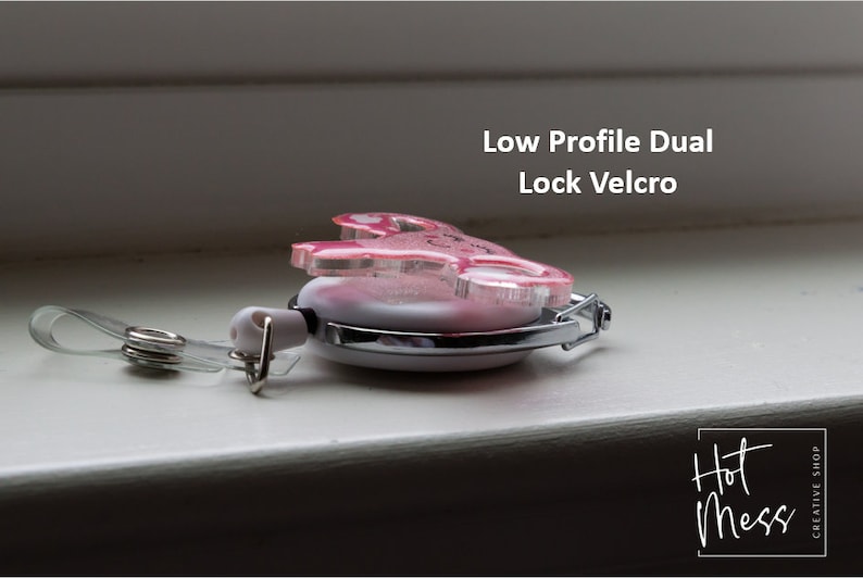 May include: A pink and white retractable badge holder with a low profile dual lock velcro closure. The holder features a pink glitter design with a heart and the text "Hot Mess" in black script. The holder is attached to a clear plastic strap with a metal clip.