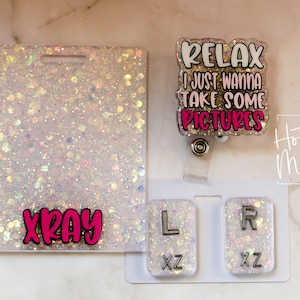 May include: A set of glittery white accessories, including an XRAY badge holder, a badge reel with the text "RELAX I JUST WANNA TAKE SOME PICTURES", and two small lettered badges. The letters are L and R.