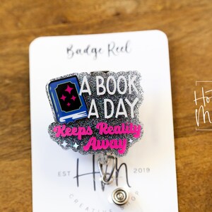 May include: A silver glitter badge reel with a blue book and pink text that reads "A book a day keeps reality away". The badge reel is attached to a white card with the text "Badge Reel" and "Hot Mess Creative Shop".