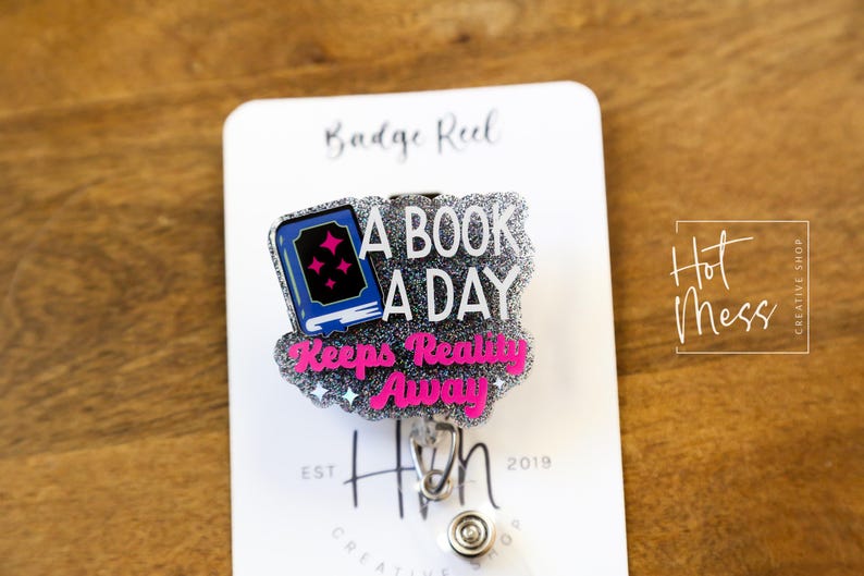 May include: A silver glitter badge reel with a blue book and pink text that reads "A book a day keeps reality away". The badge reel is attached to a white card with the text "Badge Reel" and "Hot Mess Creative Shop".