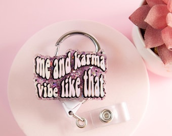 Pode incluir: Um porta-crachá com a frase "me and karma vibe like that" em texto branco com um fundo roxo brilhante. O porta-crachá tem um clipe prateado e uma alça de plástico transparente com fecho de botão. O porta-crachá está em uma superfície rosa.