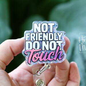 May include: A rectangular badge reel with the text "NOT FRIENDLY DO NOT TOUCH" in white and pink lettering. The badge has a glittery, iridescent border and a clear plastic reel clip.