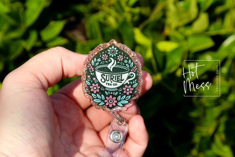 May include: A pink and green glitter badge holder with a floral design and a teacup with the text "Suriel Tea Co."
