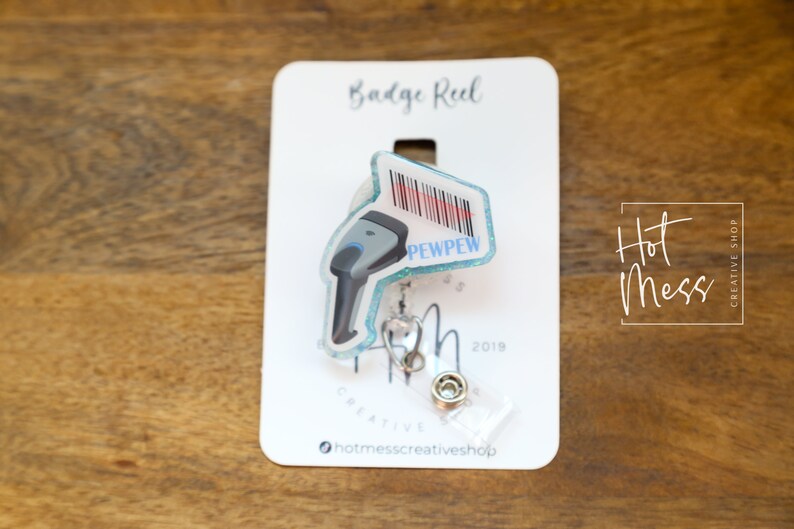 May include: A blue and white badge reel with a barcode scanner design. The scanner has the text "PEWPEW" on it. The badge reel is on a white card with the text "Badge Reel" and "Hot Mess Creative Shop" printed on it.