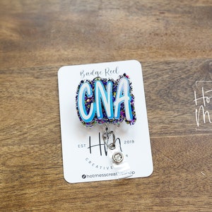 May include: A white card with the text "Badge Reel" and "Hot Mess Creative Shop" on it. A blue, white, and purple glitter badge reel with the text "CNA" is attached to the card. The badge reel is attached to a silver retractable badge holder.