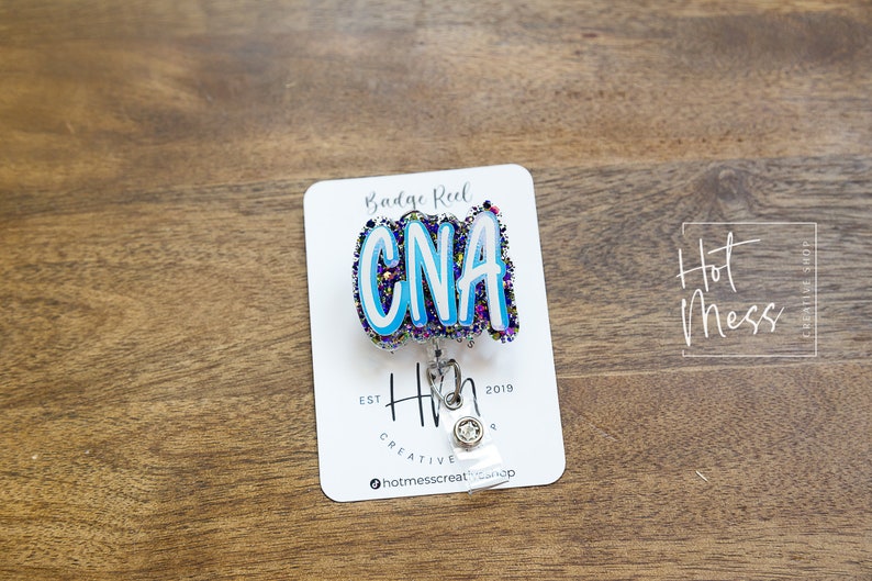 May include: A white card with the text "Badge Reel" and "Hot Mess Creative Shop" on it. A blue, white, and purple glitter badge reel with the text "CNA" is attached to the card. The badge reel is attached to a silver retractable badge holder.