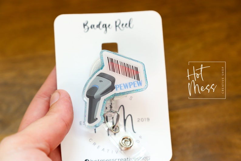 May include: A blue and white badge reel with a barcode and the text "PEWPEW" on it. The reel is attached to a white card with the text "Badge Reel" and "Hot Mess Creative Shop".