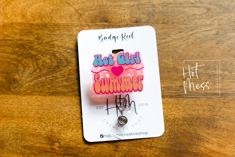 Pode incluir: Carretel de crach&aacute; de glitter rosa com um design "Hot Girl Summer" azul e rosa. O carretel &eacute; preso a um clipe prateado e tem um cart&atilde;o branco com o texto "Hot Mess Creative Shop" e "Badge Reel" impresso nele.