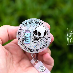 May include: A round, glittery badge reel with the text "Fueled by Energy Drinks & Anxiety." The design features a skeleton hand holding a can and a skull. The badge reel is held by a person.