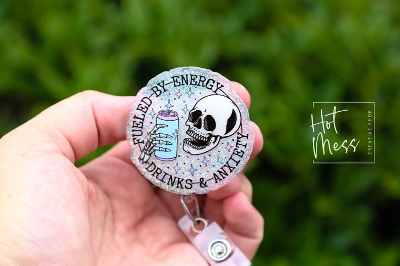 May include: A round, glittery badge reel with the text "Fueled by Energy Drinks & Anxiety." The design features a skeleton hand holding a can and a skull. The badge reel is held by a person.