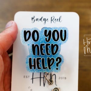 May include: A badge reel with the text "DO YOU NEED HELP?" in black lettering on a light blue glitter background. The badge reel is attached to a white card with the words "Badge Reel" at the top. The words "Hot Mess" are in a box on the right.