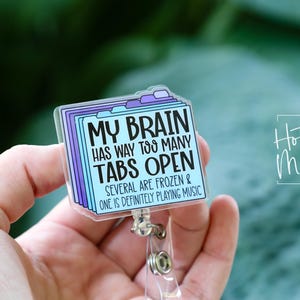 May include: A clear acrylic badge reel with a design of stacked file folders in shades of blue and purple. The text reads "MY BRAIN HAS WAY TOO MANY TABS OPEN." The badge reel has a silver clip and a clear plastic strap.