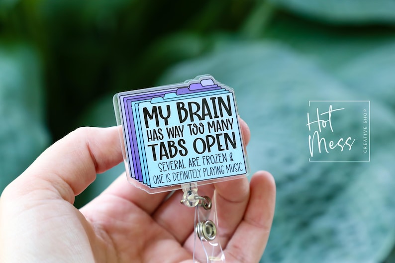 May include: A clear acrylic badge reel with a design of stacked file folders in shades of blue and purple. The text reads "MY BRAIN HAS WAY TOO MANY TABS OPEN." The badge reel has a silver clip and a clear plastic strap.