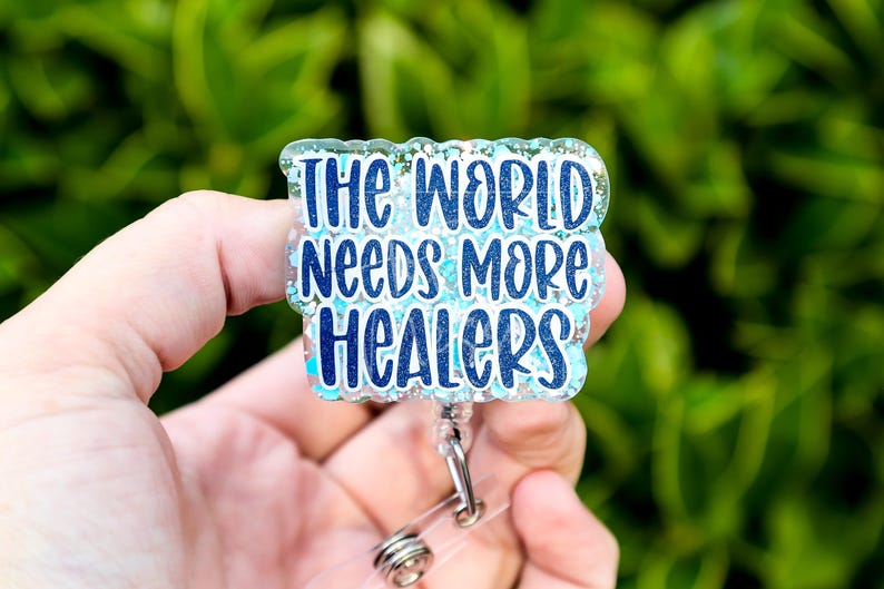 May include: A badge reel with the text "The World Needs More Healers" in blue script. The badge has a clear, glittery border and a silver retractable clip. The background is a blurred green.