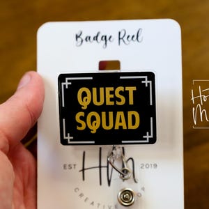 May include: A black and gold badge reel displaying the words "QUEST SQUAD" in gold. The rectangular badge reel has a white border and is attached to a white card labelled "Badge Reel". The card also features the "Hot Mess Creative Shop" logo.