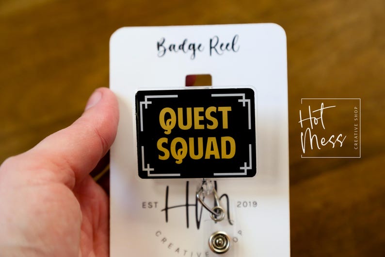 May include: A black and gold badge reel with the words "QUEST SQUAD" in gold lettering. The badge reel is rectangular with a white border and is attached to a white card that reads "Badge Reel". The card also has the "Hot Mess Creative Shop" logo.