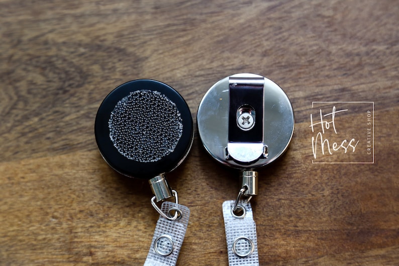 May include: Two retractable badge reels, one black and one silver, with a clip on the back and a white strap. The black reel has a textured surface and the silver reel has a smooth surface.