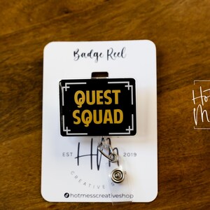May include: A black and gold badge reel with the words "QUEST SQUAD" in gold lettering. The reel is attached to a white card with the text "Badge Reel" and "Hot Mess Creative Shop". The background is a wooden surface.