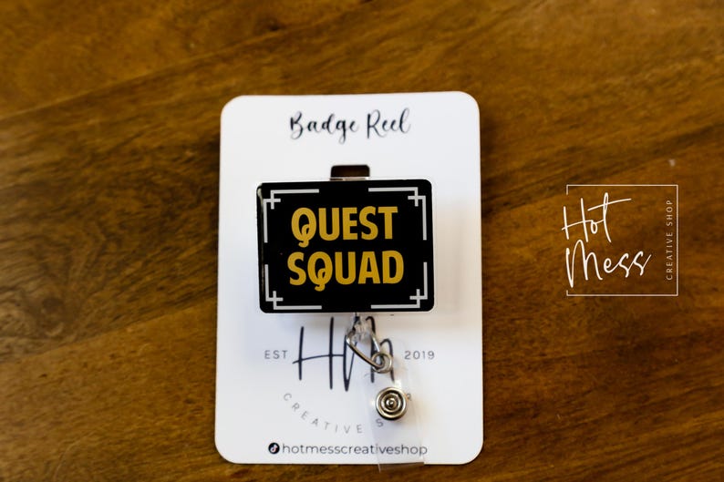 May include: A black and gold badge reel with the words "QUEST SQUAD" in gold lettering. The reel is attached to a white card with the text "Badge Reel" and "Hot Mess Creative Shop". The background is a wooden surface.
