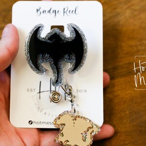Puede incluir: Un carrete de insignia de purpurina plateada con un dise&ntilde;o de drag&oacute;n negro de purpurina. El carrete est&aacute; unido a un amuleto de purpurina dorada. La tarjeta dice "Badge Reel" y "Hot Mess Creative Shop".