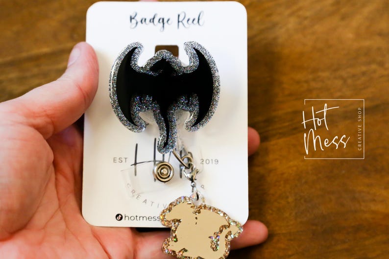 Puede incluir: Un carrete de insignia de purpurina plateada con un dise&ntilde;o de drag&oacute;n negro de purpurina. El carrete est&aacute; unido a un amuleto de purpurina dorada. La tarjeta dice "Badge Reel" y "Hot Mess Creative Shop".