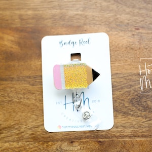 May include: A yellow and pink glitter pencil badge reel with a silver retractable badge holder. The card reads "Badge Reel" and "Hot Mess Creative Shop" with the text "Est. 2019".