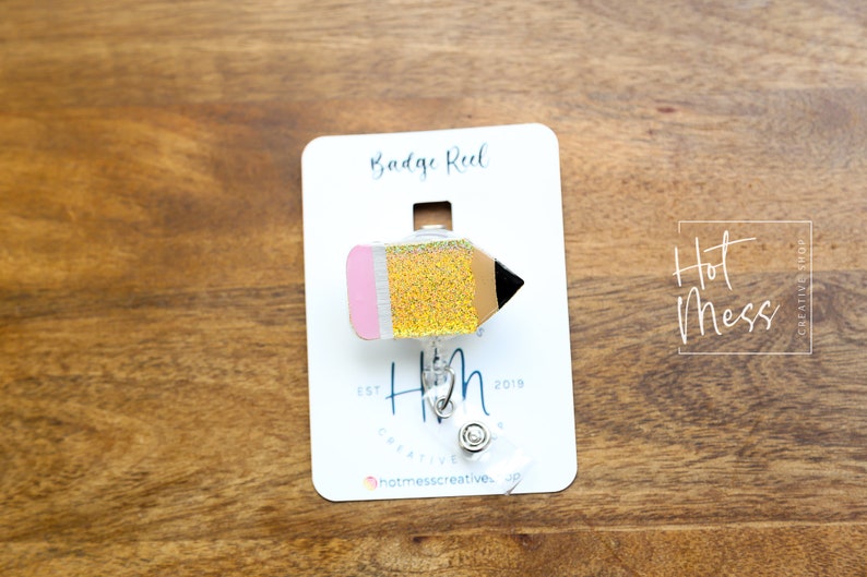 May include: A yellow and pink glitter pencil badge reel with a silver retractable badge holder. The card reads "Badge Reel" and "Hot Mess Creative Shop" with the text "Est. 2019".